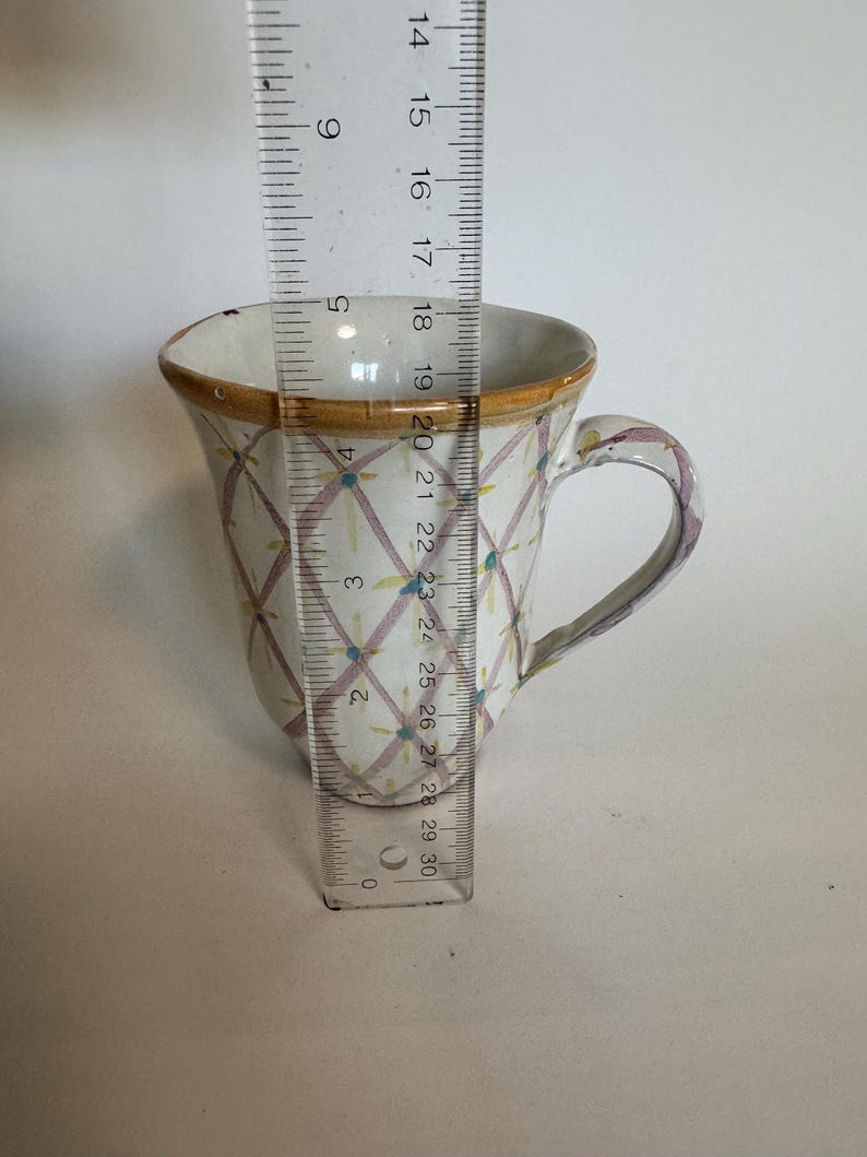 May include: A white ceramic mug with a decorative pattern of intersecting lines and small blue and pink dots. The rim is trimmed with a gold-colored band. The mug is approximately 4 inches tall.
