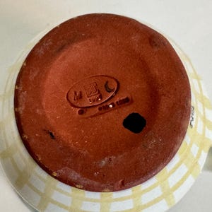 Op de afbeelding: Een keramische mok met een lichtblauw en geel geruit patroon aan de buitenkant. De bodem van de mok is roodbruin met stempelmarkeringen. De mok heeft een oor en een witte binnenkant.