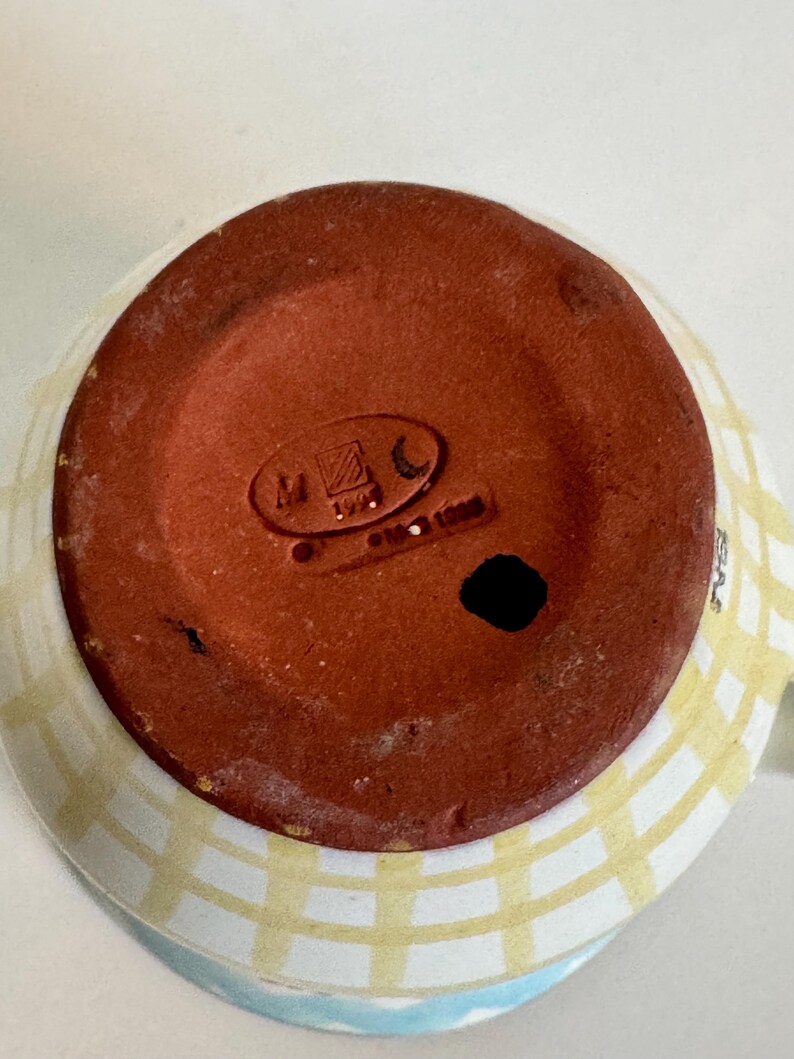 Op de afbeelding: Een keramische mok met een lichtblauw en geel geruit patroon aan de buitenkant. De bodem van de mok is roodbruin met stempelmarkeringen. De mok heeft een oor en een witte binnenkant.