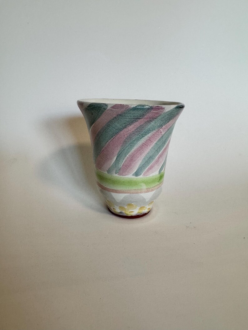 Op de afbeelding: Een keramische vaas met een uitlopende bovenkant, met diagonale groene en roze strepen. De vaas heeft een groene en gele band bij de basis, met een decoratief patroon en een rode basis.