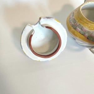 K&ouml;nnte beinhalten: Eine Keramik-Teekanne mit Deckel. Die Teekanne ist haupts&auml;chlich gelb mit einem karierten Muster in Rosa, Gr&uuml;n und Grau. Der Deckel ist wei&szlig; mit einem braunen Rand und einem kleinen Griff. Die Teekanne steht auf einer wei&szlig;en Oberfl&auml;che.