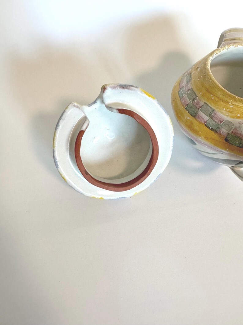 K&ouml;nnte beinhalten: Eine Keramik-Teekanne mit Deckel. Die Teekanne ist haupts&auml;chlich gelb mit einem karierten Muster in Rosa, Gr&uuml;n und Grau. Der Deckel ist wei&szlig; mit einem braunen Rand und einem kleinen Griff. Die Teekanne steht auf einer wei&szlig;en Oberfl&auml;che.