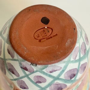 May include: A ceramic mug with a colorful, hand-painted design. The bottom of the mug is a reddish-brown color with a stamp that reads "1993". The mug features a pattern of purple ovals and green lines, with a pink and white base.