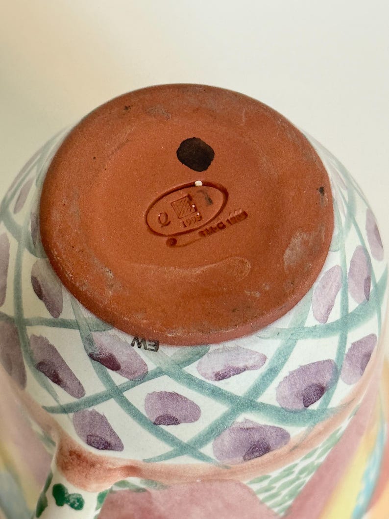 May include: A ceramic mug with a colorful, hand-painted design. The bottom of the mug is a reddish-brown color with a stamp that reads "1993". The mug features a pattern of purple ovals and green lines, with a pink and white base.
