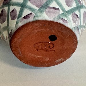 May include: A ceramic vase with a rounded base and a patterned exterior. The design features a white background with a green crisscross pattern and purple floral accents. The base is a reddish-brown color and has a small hole.