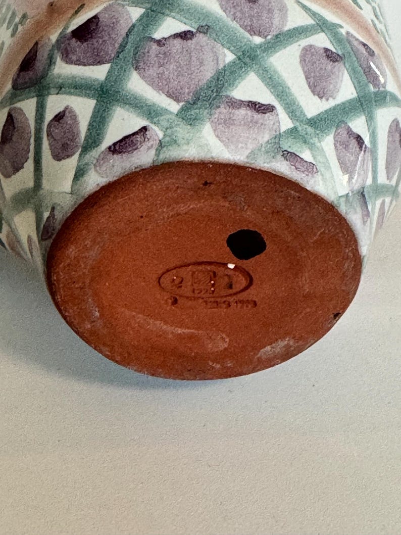 May include: A ceramic vase with a rounded base and a patterned exterior. The design features a white background with a green crisscross pattern and purple floral accents. The base is a reddish-brown color and has a small hole.