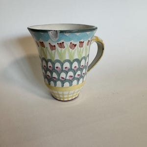 Op de afbeelding: Een keramische mok met een uitlopende bovenkant en een gele handgreep. De mok heeft een handgeschilderd ontwerp met een blauwe rand, rode tulpachtige bloemen en een herhalend patroon van groene en roze vormen. De basis heeft een geel rasterpatroon.