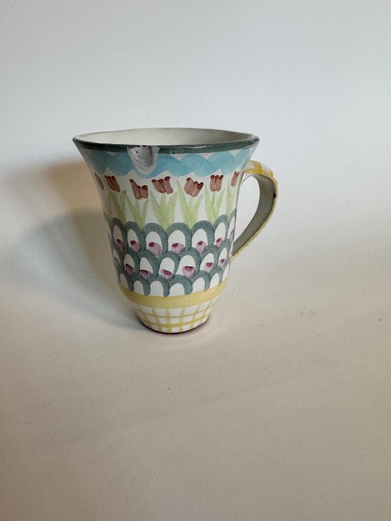 Op de afbeelding: Een keramische mok met een uitlopende bovenkant en een gele handgreep. De mok heeft een handgeschilderd ontwerp met een blauwe rand, rode tulpachtige bloemen en een herhalend patroon van groene en roze vormen. De basis heeft een geel rasterpatroon.