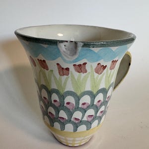 Op de afbeelding: Een keramische mok met een bloemenontwerp. De mok heeft een witte binnenkant, een lichtblauwe golfband en een groene rand. De buitenkant is versierd met rode bloemen, groene bladeren en een patroon van turquoise en witte vormen. Het handvat is geel.