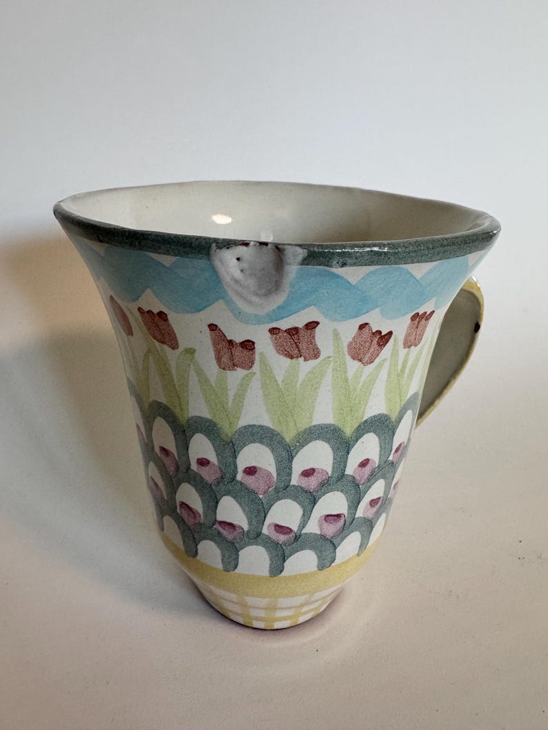 Op de afbeelding: Een keramische mok met een bloemenontwerp. De mok heeft een witte binnenkant, een lichtblauwe golfband en een groene rand. De buitenkant is versierd met rode bloemen, groene bladeren en een patroon van turquoise en witte vormen. Het handvat is geel.