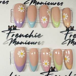 May include: A set of French manicured press-on nails with a daisy design. The nails have a nude base with pastel tips in purple, blue, pink, green, and peach. Each nail features a white daisy with a yellow centre. The text "Frenchie Manicures" is above and below the nails.