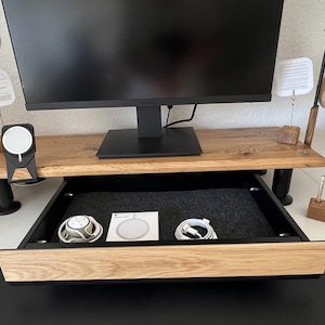 May include: A wooden desk with a monitor, a drawer, and accessories. The desk has a natural wood finish and a black drawer. The drawer contains a charging pad, cables, and a charging station. A pair of headphones hangs on the side.