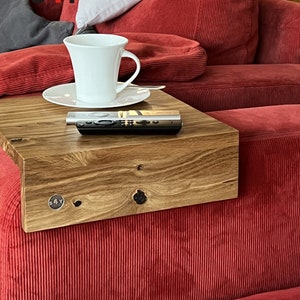 Könnte beinhalten: Ein Holzarmlehnentablett mit einer Tasse und Untertasse, einer Fernbedienung und einem kleinen Metallschild mit dem Text "s4y". Das Tablett ist an einem roten Cord-Sofa befestigt.