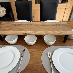 Peut inclure: Un plateau de service en bois avec des pieds noirs, posé sur une table en bois. Des bols blancs sont placés sous le plateau. Des assiettes blanches, des couverts et des sets de table gris sont disposés sur la table.