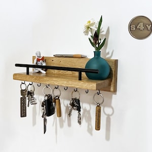 Könnte beinhalten: Schlüsselhalter aus Holz mit einer schwarzen Metallstange und Haken. Der Halter hat ein kleines Regal mit einer Vase mit weißen Blumen und einer kleinen Figur. Der Halter ist an einer weißen Wand mit einem Metallschild befestigt, auf dem "S4Y" steht.