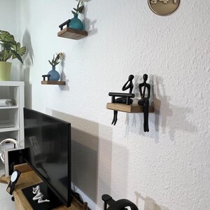 Może przedstawiać: Nowoczesny salon z drewnianymi półkami. Dwie półki prezentują małe niebieskie wazony z białymi kwiatami. Trzecia półka prezentuje czarną metalową rzeźbę dwóch postaci. Widoczna jest również drewniana tablica z napisem "s4y".