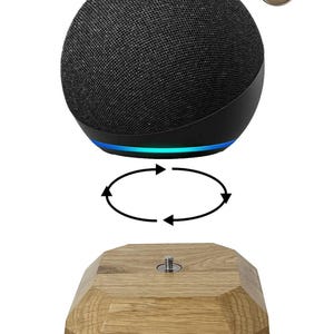 Op de afbeelding: Een zwarte slimme luidspreker met een stoffen hoes en een blauw licht aan de basis. Het is gepositioneerd boven een houten, geometrische standaard. Een klein, cirkelvormig embleem met de letters "S4Y" staat in de rechterbovenhoek.