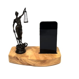Puede incluir: Un soporte de teléfono de madera con una pequeña estatua de bronce de la diosa de la justicia sosteniendo una balanza y una espada. El soporte tiene un hueco para un teléfono y una pequeña plataforma para la estatua.