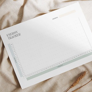May include: A white energy tracker sheet with the title "ENERGY TRACKER" in a stylish font. The sheet has a grid for tracking energy levels, with a light green and beige color scheme. The sheet is placed on a beige fabric.