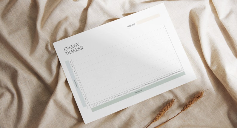 May include: A white energy tracker sheet with the title "ENERGY TRACKER" in a stylish font. The sheet has a grid for tracking energy levels, with a light green and beige color scheme. The sheet is placed on a beige fabric.
