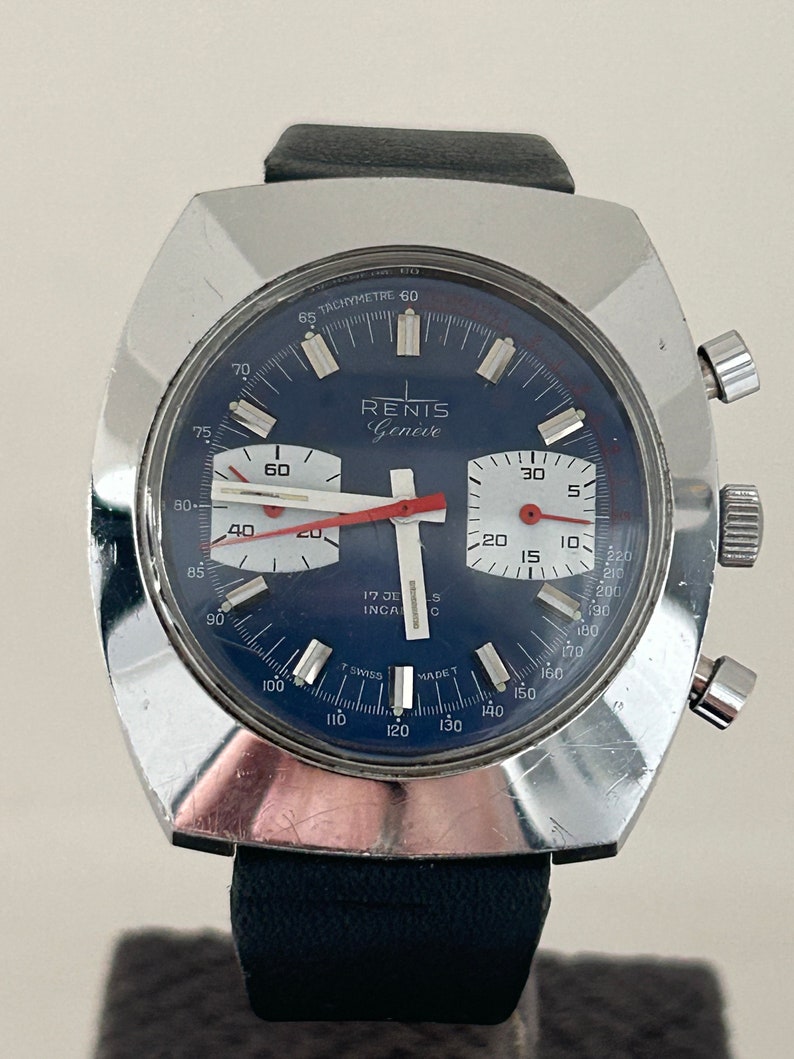 May include: A silver chronograph wristwatch with a blue face and red accents. The watch face has the text "RENIS Gen&egrave;ve" and "17 JE INCA C" printed on it. The watch has a black leather band.