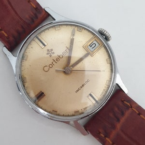 May include: A gold-toned wristwatch with a brown leather band. The watch face has black markings and the text "Cortebert" and "Incabloc" printed on it. The watch has a date window at the 3 o'clock position.