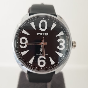 May include: A black and silver wristwatch with a black leather strap. The watch face has large white numbers and the word "PAKEТА" in the centre. The watch face also has the text "СДЕЛАНО В СССР" at the bottom.