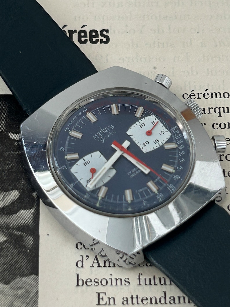 May include: A silver chronograph wristwatch with a black face and red accents. The watch has a square case and a black leather band. The watch face features the text "RENIS Genive" and a tachymeter scale.