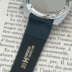 May include: Close-up of the back of a silver wristwatch with a black leather band. The watch face is marked with "SWISS MADE" and "ANTIMAGNETIC" and has the number "01007 V" printed on it. The band is labeled "201 HIRSCH 1.Quality Calf".