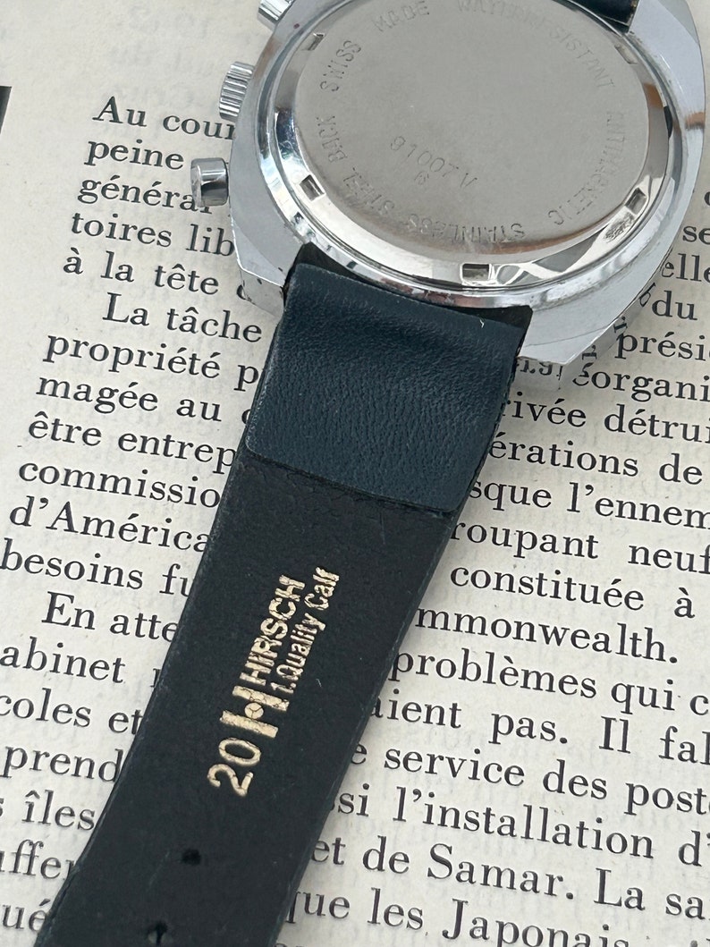 May include: Close-up of the back of a silver wristwatch with a black leather band. The watch face is marked with "SWISS MADE" and "ANTIMAGNETIC" and has the number "01007 V" printed on it. The band is labeled "201 HIRSCH 1.Quality Calf".