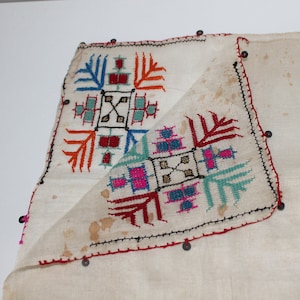 Könnte beinhalten: Off-white Textil mit farbenfrohen, gestickten geometrischen Mustern. Die Stickerei zeigt rote, blaue, orangefarbene und türkisfarbene Designs. Die Kanten sind mit roten Nähten und kleinen, dunklen Perlen versehen.