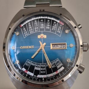 Può includere: Un orologio Orient argentato con quadrante blu e display del calendario. L'orologio ha un cinturino argentato e una finestra della data che mostra il giorno della settimana e la data. L'orologio ha anche un display del giorno e del mese.