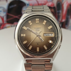 Pode incluir: Um relógio Seiko 5 Automatic prateado com um mostrador marrom e detalhes dourados. O relógio tem uma janela de data que mostra "TUE 1" e uma janela de dia da semana que mostra "TUE". O relógio tem uma pulseira de metal prateada.