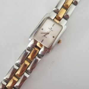 May include: A rectangular Junghans Quartz watch with a silver and gold-tone metal band. The watch face is silver with black hands and markers. The band alternates silver and gold-tone links.