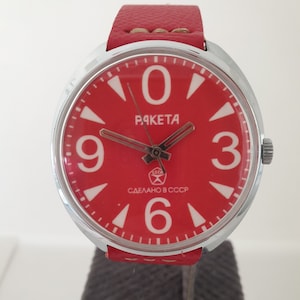 May include: A red wristwatch with a white face and black hands. The watch face has large white numbers and the words "PAKEТА" and "СДЕЛАНО В СССР".