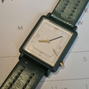 May include: A vintage Seiko watch with a square, off-white face and gold hands. The watch has a dark green leather band. The watch face displays the numbers 3, 6, 9, and 12. The watch is on a white background.
