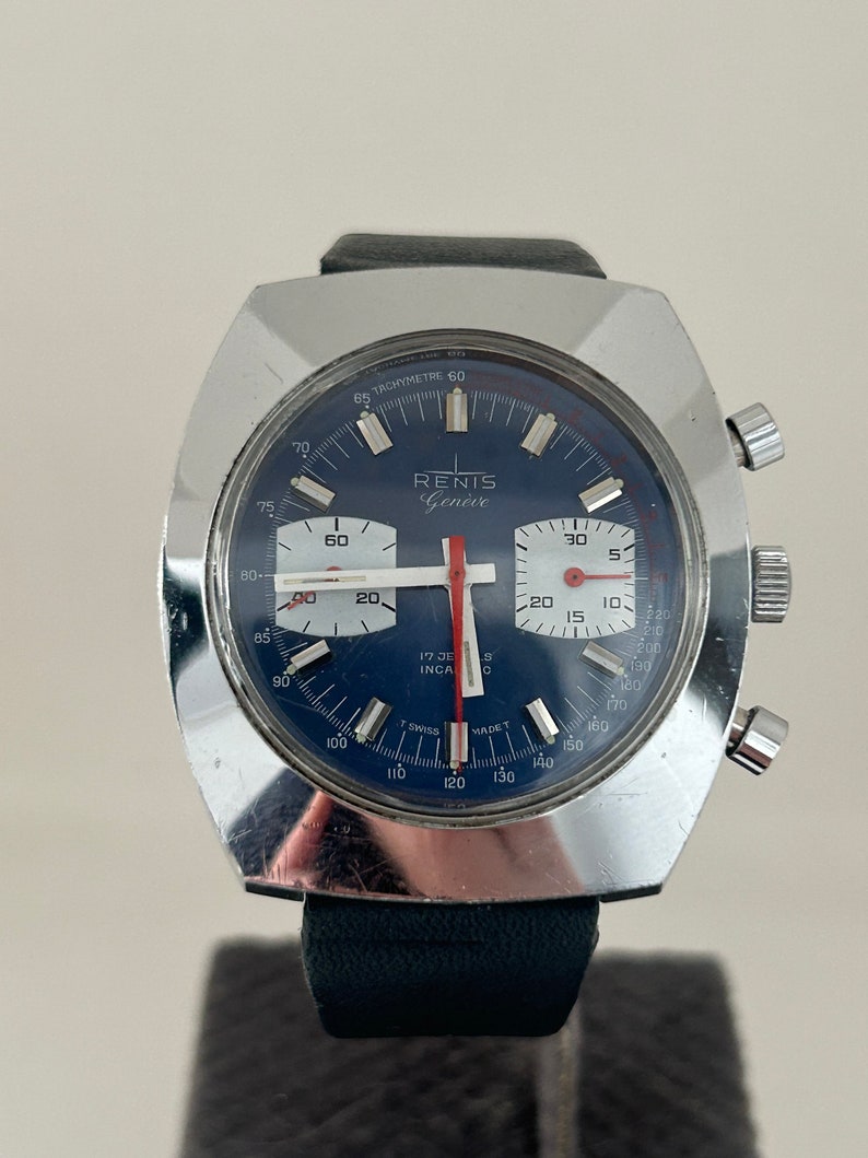 May include: A silver chronograph wristwatch with a blue face and red accents. The watch face features the text "RENIS Gen&egrave;ve" and "TACHYMETRE 60".