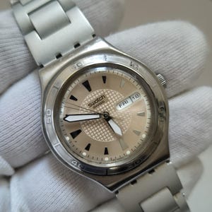 May include: A silver Swatch watch with a white face and black hands. The watch has a date window that displays "FRI 18".