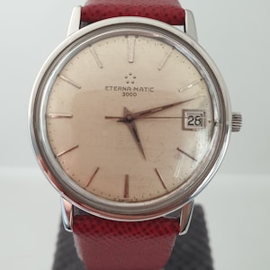 Puede incluir: Un reloj de pulsera plateado con una esfera blanca y una correa de cuero roja. La esfera del reloj tiene marcadores de hora romanos negros, una ventana de fecha a las 3 en punto y el texto "ETERNA-MATIC 3000".