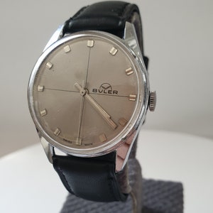 Könnte beinhalten: Eine silberne Armbanduhr mit einem schwarzen Lederarmband. Das Zifferblatt der Uhr ist silberfarben mit schwarzen Markierungen und dem Wort "BULER" in Schwarz gedruckt.