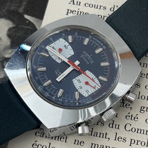 May include: A silver chronograph wristwatch with a blue face and white markings. The watch has a tachymeter scale around the outer edge of the face. The watch is on a black leather strap.
