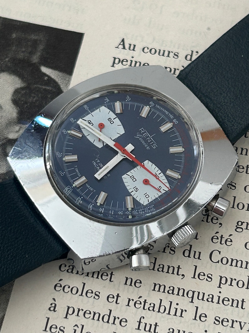 May include: A silver chronograph wristwatch with a blue face and white markings. The watch has a tachymeter scale around the outer edge of the face. The watch is on a black leather strap.