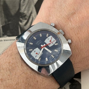 May include: A silver chronograph wristwatch with a blue face and red accents. The watch has a square case and a black leather strap. The watch face has the text "RENIS Geneve" and "17 Jewels Incabloc".