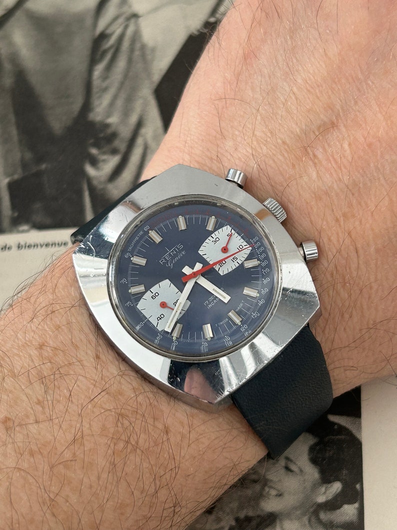 May include: A silver chronograph wristwatch with a blue face and red accents. The watch has a square case and a black leather strap. The watch face has the text "RENIS Geneve" and "17 Jewels Incabloc".
