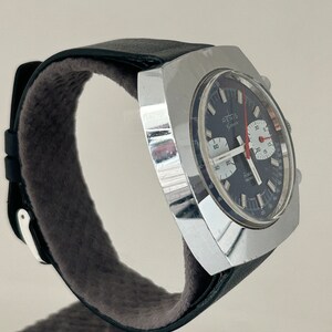 May include: A silver square-shaped chronograph wristwatch with a black face and red second hand. The watch has a black leather band with a white stitching detail.