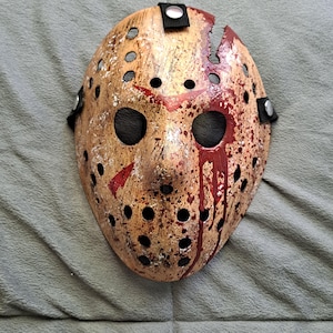 Op de afbeelding: Een hockeymasker met een verweerde, beige afwerking met versleten verfspatten en gesimuleerde bloeddruppels. Het masker heeft meerdere ronde ventilatiegaten, twee oogopeningen, een scheur aan de bovenkant en zwarte elastische banden.
