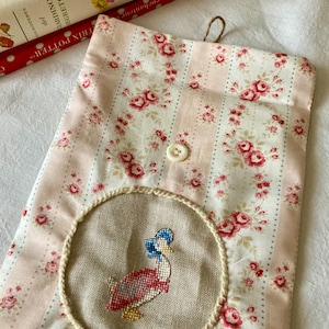 Può includere: Una pochette in tessuto a righe rosa e bianche con un motivo floreale. La pochette ha una toppa rotonda ricamata con un'anatra che indossa un berretto blu. La pochette ha un bottone bianco e uno spago marrone.