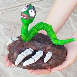 May include: A green worm with a toothy grin and large eyes is perched on a brown rock with white claws. The worm is made of clay and has a green and white striped body.
