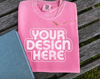 May include: Pink t-shirt with the text "Your Design Here" in white. The t-shirt is laying on a wooden bench with a blue notebook next to it.