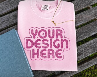 May include: A pink t-shirt with a distressed, faded look. The shirt has a white tag with the text "COMFORT COLORS" and the text "YOUR DESIGN HERE" in a pink, distressed font.